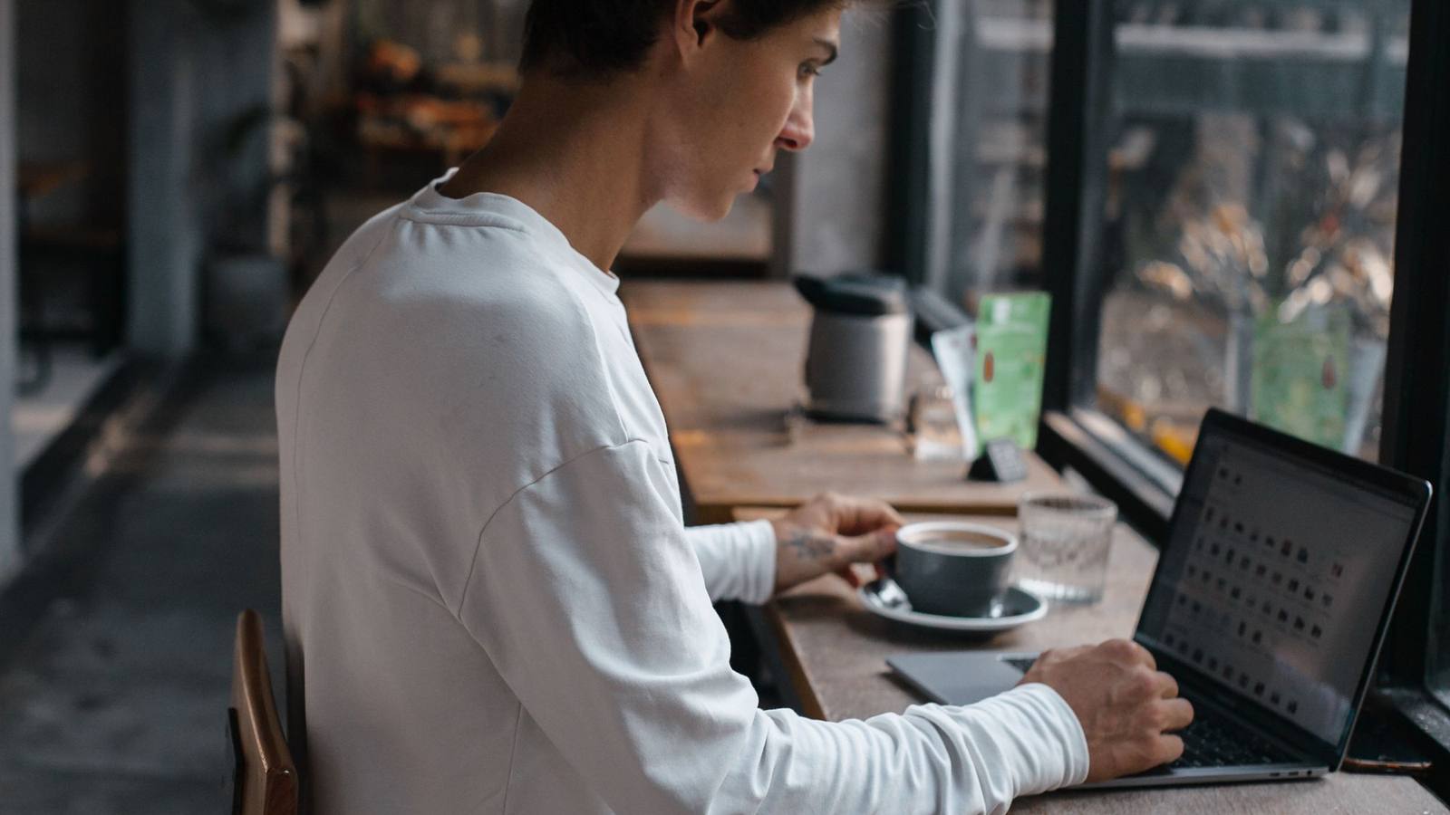 A young man working on a laptop at a café table with a coffee beside him.